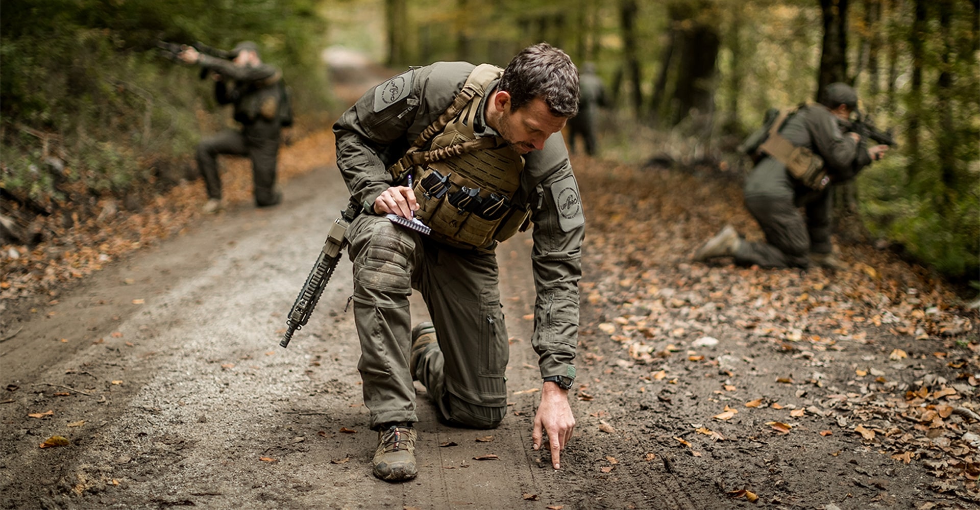 Three operators kneeling on muddy forest terrain, following a trail and securing the perimeter while searching for signs.